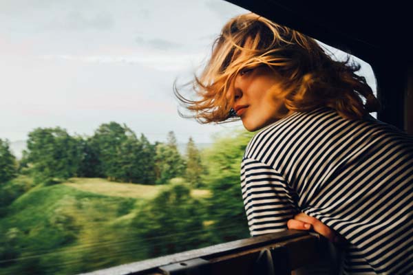 Blonde Frau schaut aus einem Fenster in der Bahn, ihre Haare fliegen im Wind